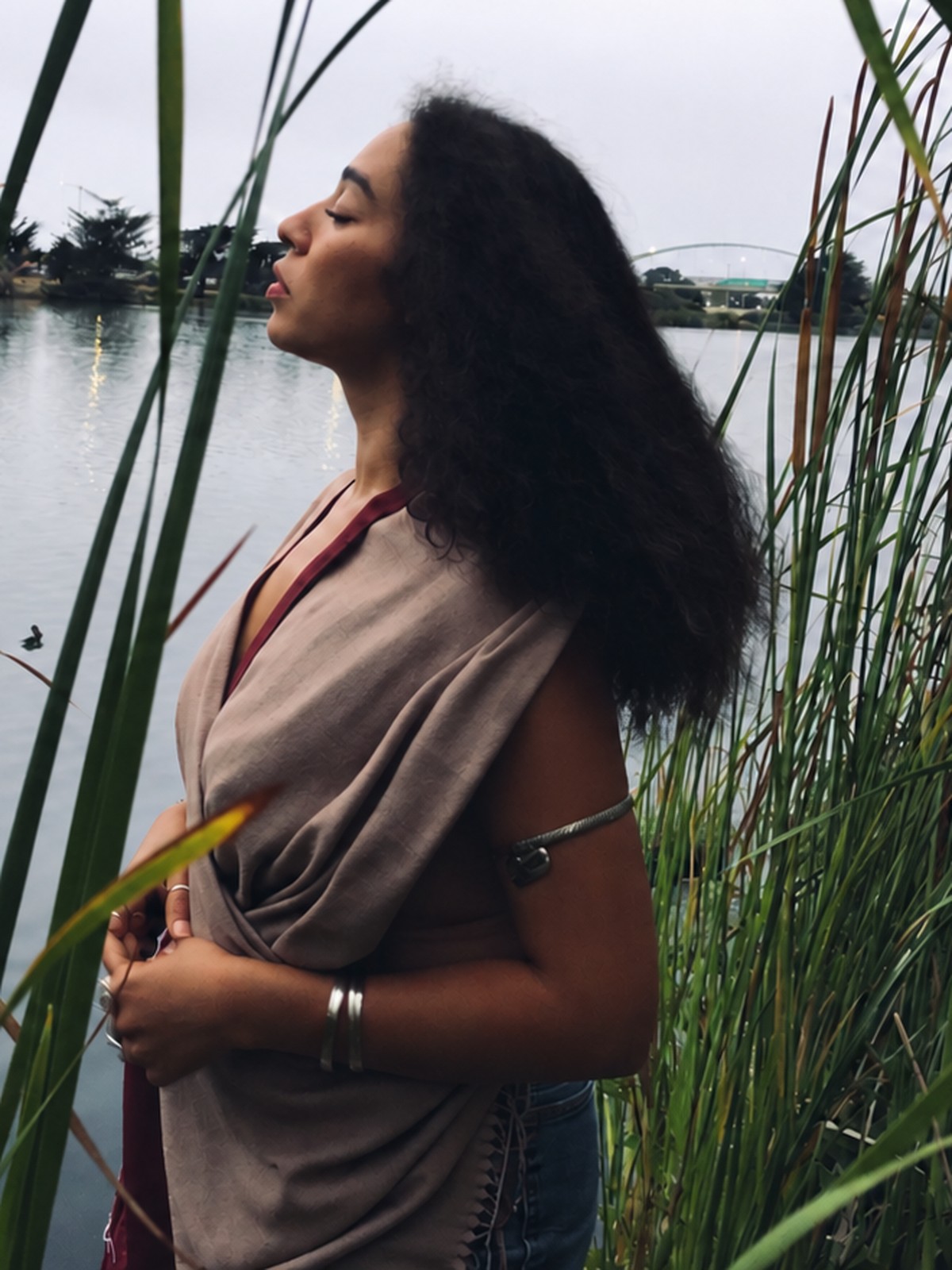 Sí in profile among tall reeds by the water, eyes closed, hair moving in the wind